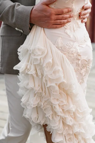 Wedding couple embrace as the groom holding bride waist, highlighting floral appliqué and ruffled tulle train on an outdoor walkway backdrop