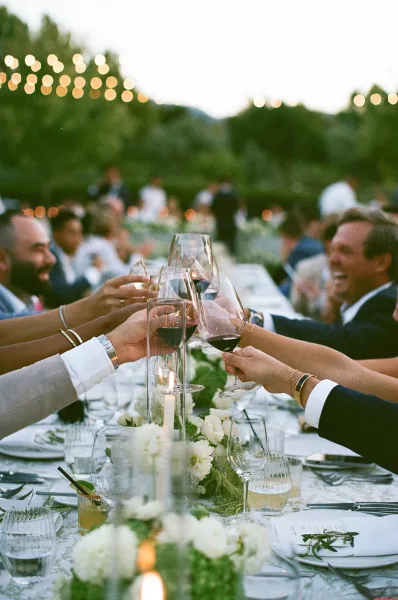 Reception toast during a wedding cheers moment as guests clink wine glasses under garden string lights at dusk along a long banquet table.