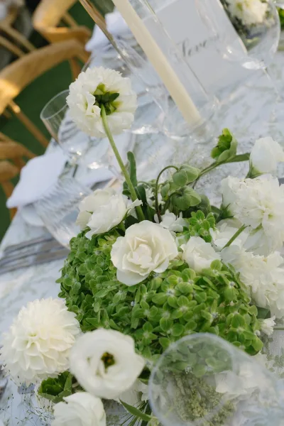 Wedding centerpiece featuring a white rose centerpiece and fresh greenery in a low arrangement, accented by taper candles on a lawn with wooden chairs
