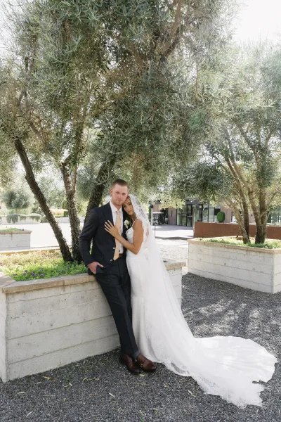 Couple portrait of bride and groom under olive trees, her lace veil draping as they embrace in a quiet gravel courtyard.