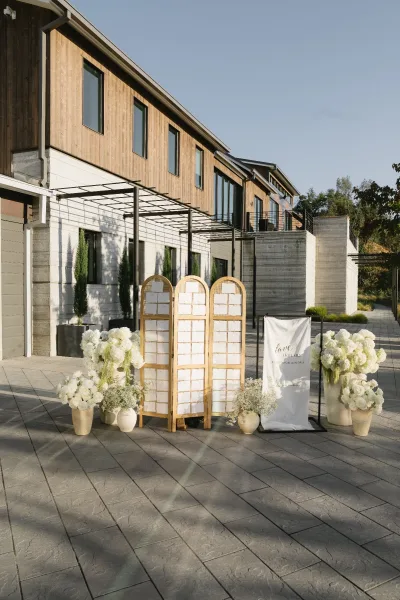 Escort card display on a rattan room divider offering chic seating chart ideas, framed by a modern building facade in a paved courtyard.