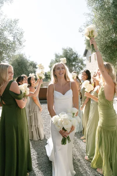 Bride with bridesmaids, sage green bridesmaids around her, holding white rose bouquets in a gravel courtyard of olive trees and a stone building.