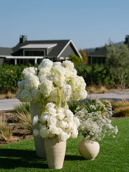 Ceremony floral arrangement featuring a hydrangea wedding arrangement in ceramic vases, set on a vineyard lawn with a farmhouse backdrop.