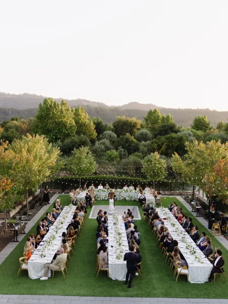 Outdoor reception layout with U-shaped reception tables under string lights, set on a lawn amid garden trees and rolling hills.