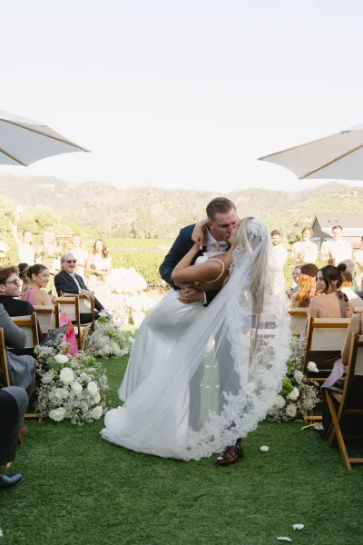 Recessional kiss as groom dips bride, her long lace veil trailing, down a vineyard aisle with mountain views under a bright sky and green lawn.