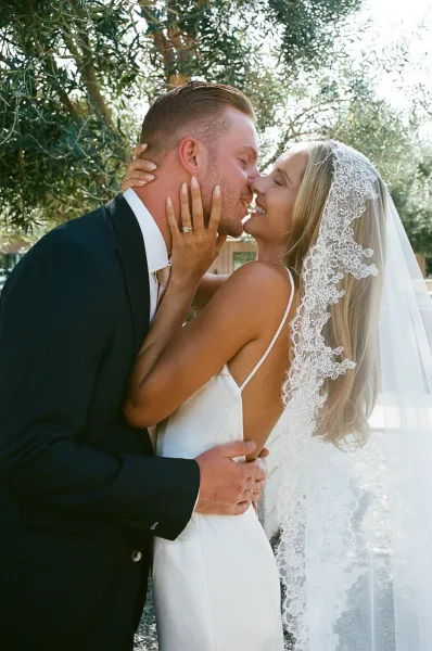 Wedding kiss portrait as a bride and groom kiss, her lace veil catching sunlight beneath trees along an outdoor path.
