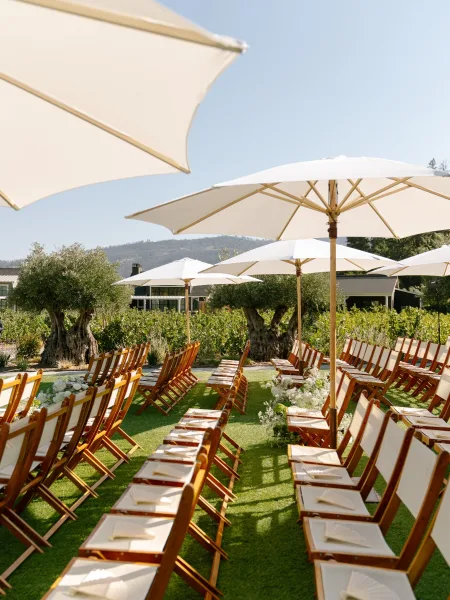 Ceremony setup featuring outdoor ceremony seating shaded by white umbrellas on a vineyard lawn, framed by rolling hills beneath a clear blue sky.