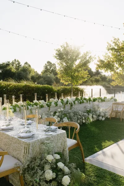 Reception tablescape for an outdoor wedding reception with taper candles, set beside a sunlit lawn and pond beneath trees and hedges.
