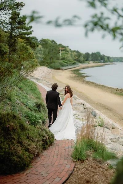 Couple portrait of a bride and groom walking along a brick path by the beach, her bridal gown trailing as waves lap and a cloudy sky frames the shore.
