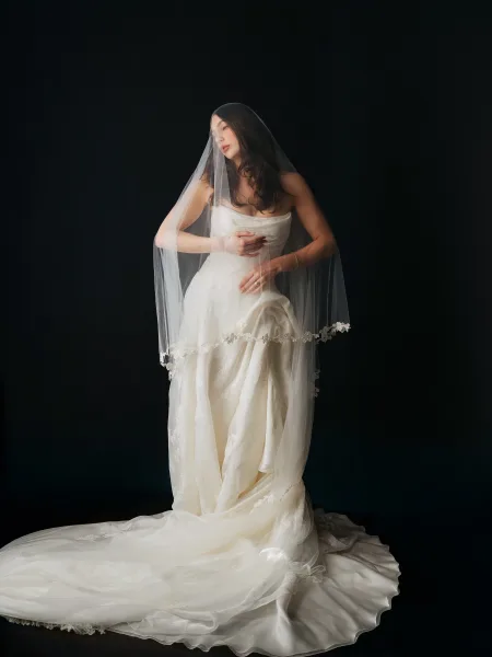 Bridal portrait of a bride with veil, softly illuminated against a black backdrop, her serene pose emphasizing elegant, timeless style.