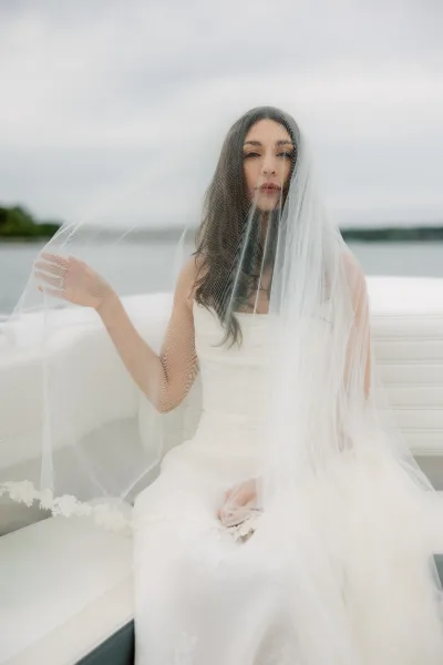 Bridal portrait of a bride with veil, seated in a boat interior as water stretches toward a cloudy sky and a faint distant shoreline.