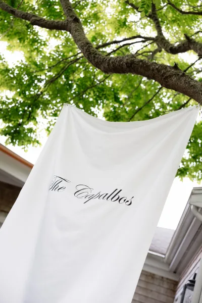 Wedding signage featuring a last name banner in elegant calligraphy lettering, hung from tree branches with green leaves near a house roof.