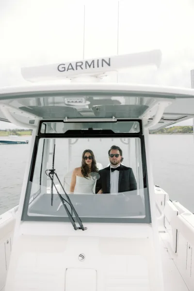 Couple portrait of a bride and groom on a boat with blue water and distant shoreline trees under a clear sky.