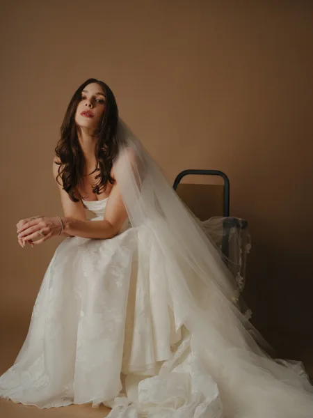 Bridal portrait of a bride in a sitting pose with a long veil softly draped over her shoulders, set against a minimal beige studio backdrop.