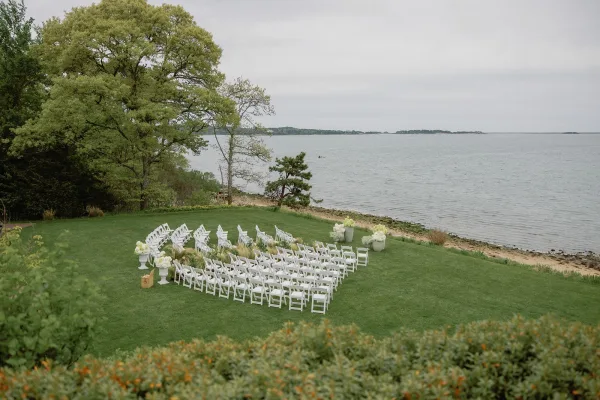 Outdoor ceremony setup with waterfront ceremony decor, rows of white folding chairs facing the water, with trees and a cloudy sky along the shoreline.