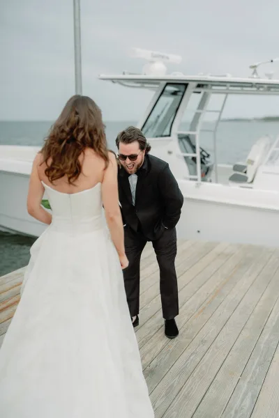 Wedding first look capturing a groom reaction photo as the bride approaches on a wooden dock, groom in sunglasses, boat and water behind.