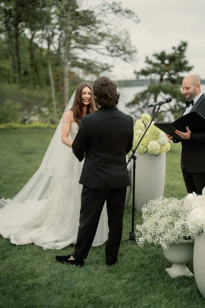 Ceremony moment as the couple share wedding vows in a lakeside garden, the bride's long veil trailing while they face the officiant under trees.