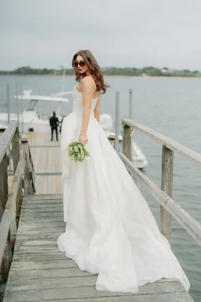 Bridal portrait of a bride on a dock, holding a lily of the valley bouquet with boats on calm water and shoreline trees under a cloudy sky.