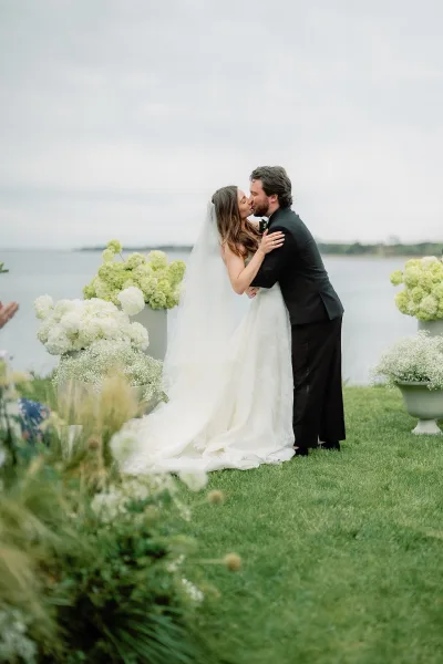 Wedding first kiss on a waterfront lawn during an outdoor ceremony moment, framed by the bride's long veil beneath a soft, cloudy sky.