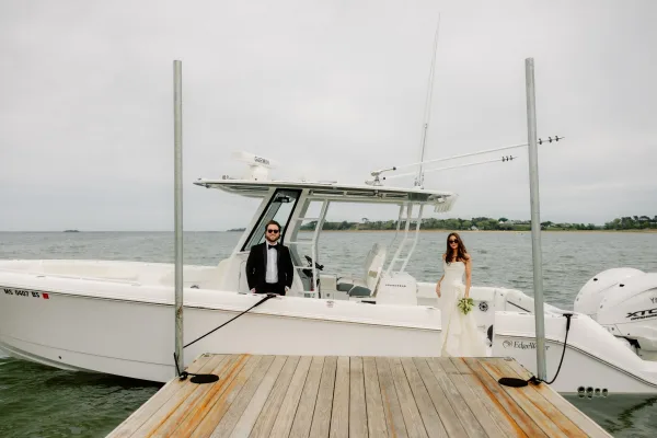 Couple portrait of bride and groom on boat, the bride holding a simple white bouquet, with dock and water beneath a cloudy sky along the shoreline.