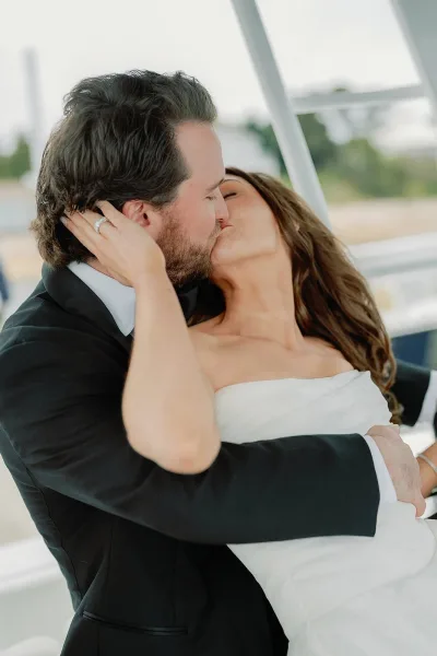 Wedding kiss portrait as the bride and groom embrace, her diamond engagement ring catching light by a white railing and soft greenery.