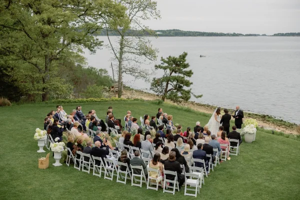 Ceremony moment at an outdoor wedding ceremony as the bride and groom stand on a waterfront lawn, framed by white florals with trees and shoreline.
