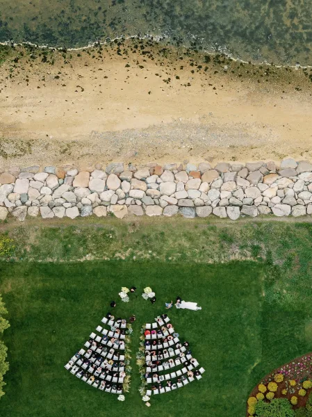 Outdoor ceremony setup with outdoor ceremony seating in a fan-shaped layout and aisle florals, set by a sandy shoreline and rock wall.