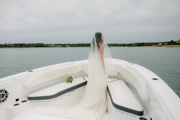 Bridal portrait of a bride on a boat, framed by rippling water and a shoreline beneath a soft, cloudy sky.