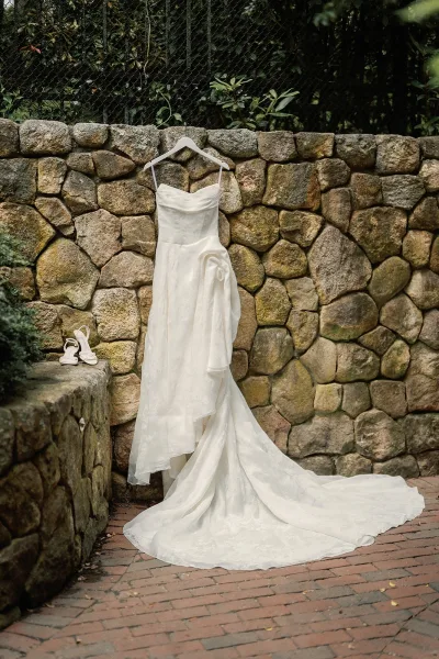 Wedding dress on hanger, displayed against a stone wall with garden greenery bordering a brick pathway.