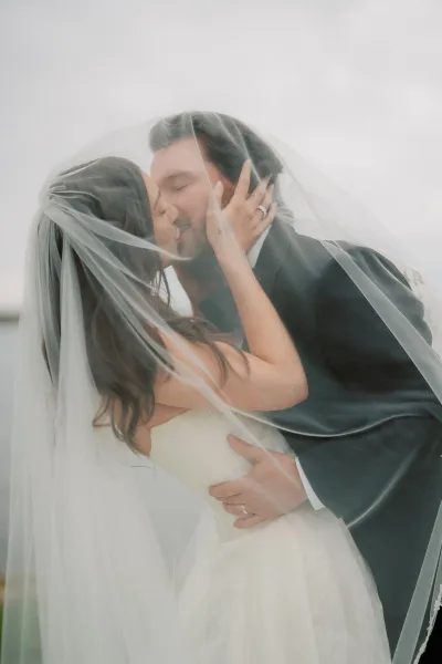 Wedding kiss portrait under the bride’s tulle veil by the water and sky, a veil kiss photo capturing an intimate, natural-light embrace.