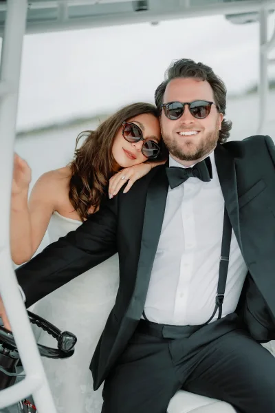 Couple portrait of a bride leaning on groom aboard a boat, beside the boat wheel, water and an overcast sky in the background.