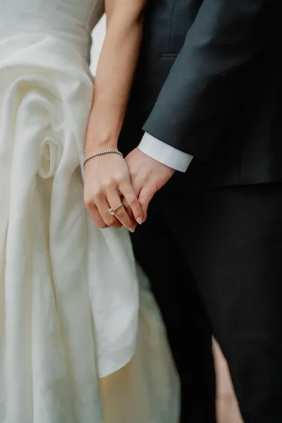 Couple holding hands in a bridal hand close-up, highlighting an engagement ring against wedding dress and dark suit fabric.