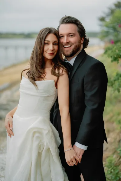 Couple portrait of a bride and groom holding hands by the waterfront, with greenery and a cloudy sky, the groom marked by a black bow tie.