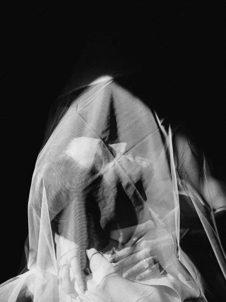 Couple portrait of a bride and groom under veil, sharing an intimate embrace, tulle framing their faces against a black backdrop with shadows.