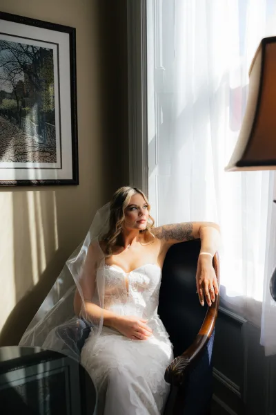 Bridal portrait of a bride sitting by window in a strapless lace wedding dress, veil draped over shoulders, showing ring in soft light