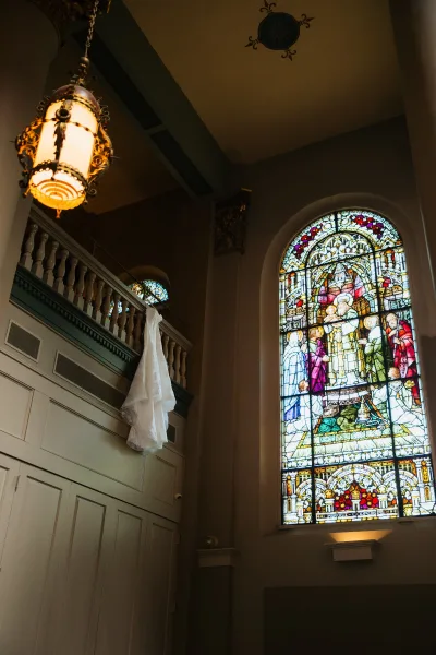 Wedding dress with lace wedding dress detail hanging from a balcony railing beside stained glass in a church interior with lantern
