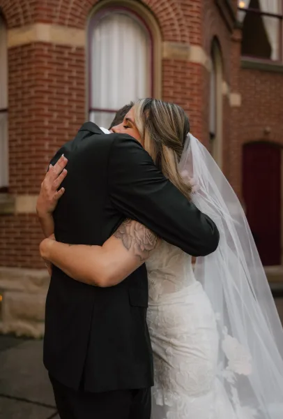 Wedding hug as bride and groom embrace, her lace dress and veil draped over a tattooed arm, beside a brick building with arched windows
