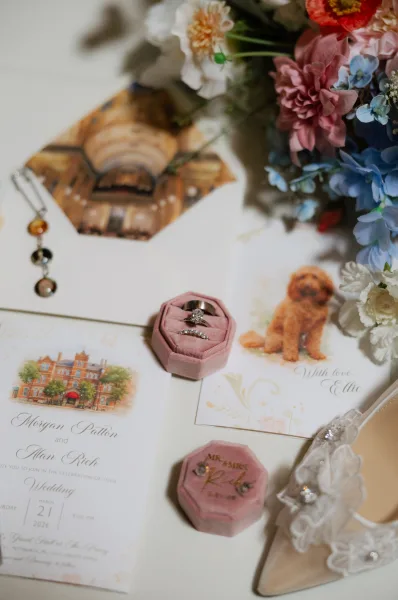 Wedding invitation suite flat lay with illustrated invites, wax seal envelope, velvet ring boxes, rings and bridal shoes on linen fabric