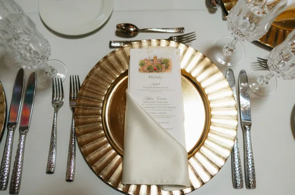 Reception place setting with a wedding tablescape featuring a gold charger plate, menu card on layered plates, and crystal glassware on white linen