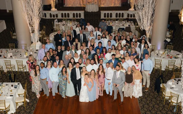 Wedding group photo of bride and groom with guests in a ballroom, seen overhead on the dance floor under string lights and tables