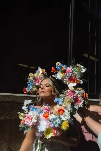 Bridal portrait of a bride with flower crown holding a colorful wedding bouquet, dramatic sunlight shadows against a dark wall by a window