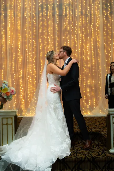 Wedding kiss as bride in lace gown and veil leans into groom in suit with glasses, under string lights and draped backdrop indoors