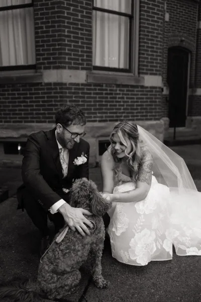 Couple portrait of newlyweds with dog, bride in lace dress and veil beside groom in suit and glasses on brick steps outdoors
