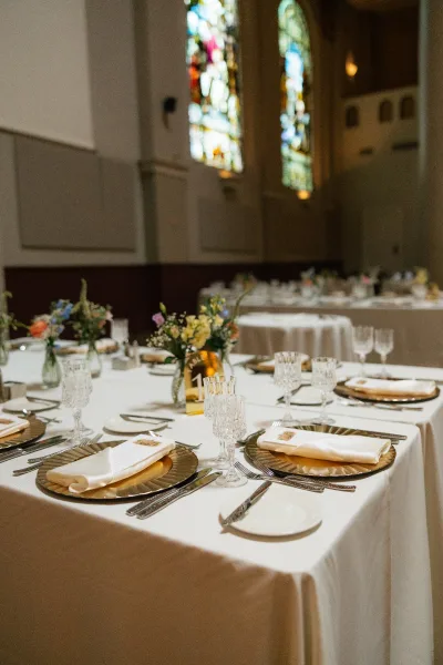 Reception tablescape wedding table setting with gold charger plates, crystal goblets, menu cards, and amber votive candles in a stained glass banquet hall