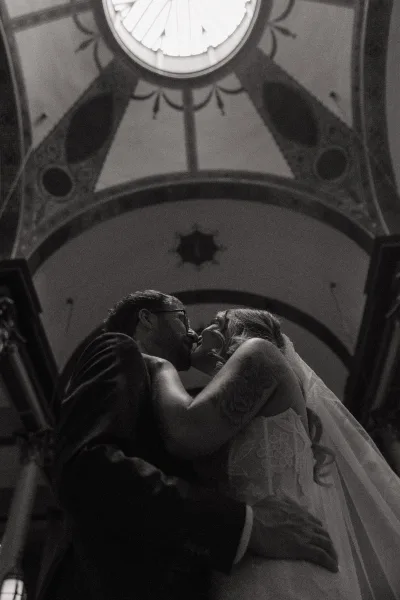 Wedding kiss beneath a dramatic church ceiling, bride’s veil and lace dress brushing the groom in glasses under an arched dome skylight