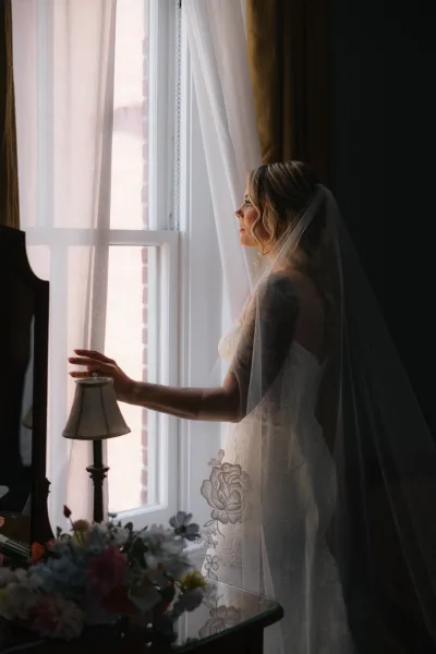 Bridal portrait of a bride by window in side profile, holding curtains in a lace wedding dress with embroidered veil in a softly lit room