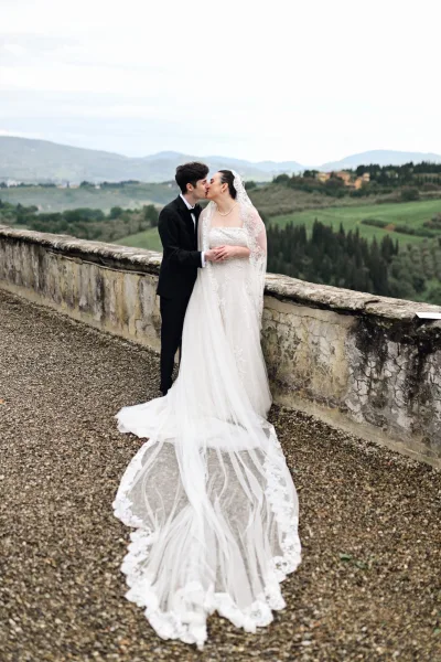 Wedding kiss portrait on a terrace, echoing a terrace wedding portrait style, the bride's cathedral veil flows before a stone wall and rolling hills.