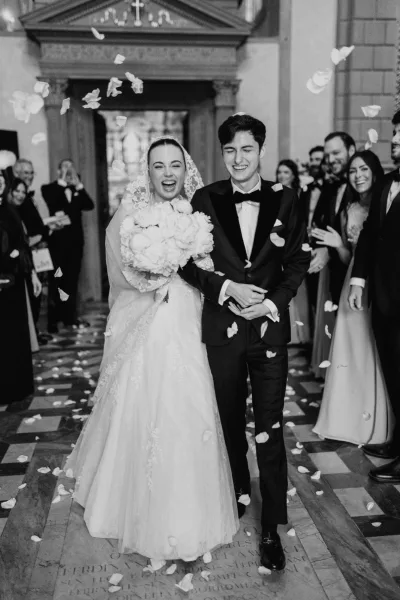 Ceremony exit during a petal toss as joyful newlyweds walk arm in arm down a stone-floored church aisle, her lace veil trailing.