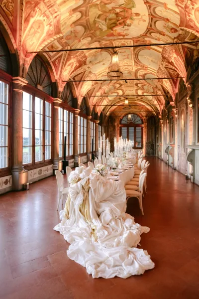 Reception tablescape with long banquet seating, lit by taper candles beneath a frescoed ceiling and arched windows in a historic hall.