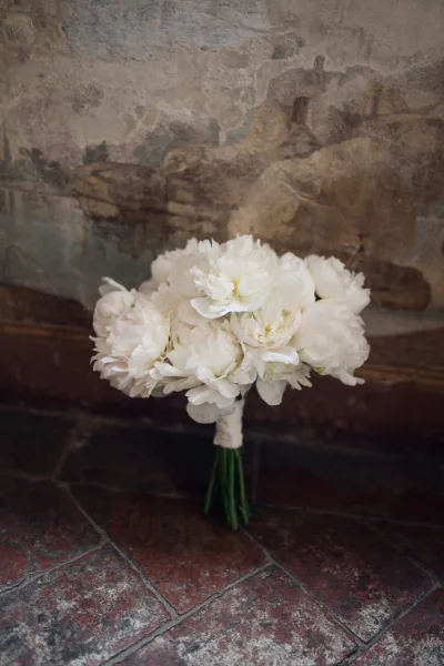Bridal bouquet, a white peony bouquet hand-tied with a ribbon, set against a textured wall and stone tile floor.